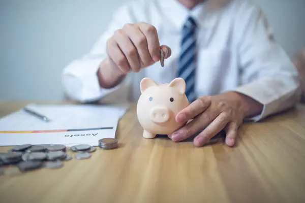 Person in a business shirt saving money in a piggy bank, with coins and financial reports on the table. Saving money business concept.