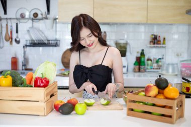 beautiful asian woman smiling preparing, cut green apple in white kitchen with plenty of fresh vegetables and fruits on the table.