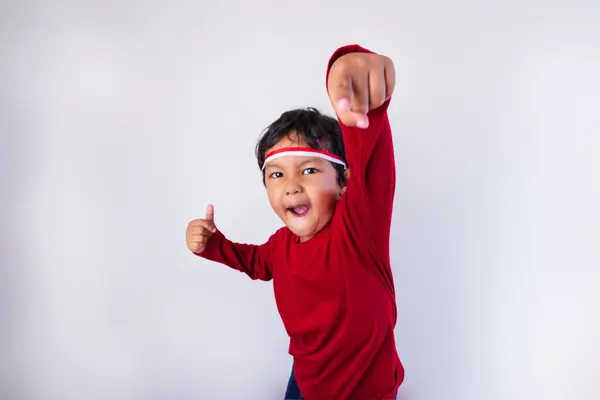 stock image A young boy wearing an Indonesian flag headband and a red shirt, jumping and pointing down while looking at the camera with a spirited expression, in front of a white background, celebrating Indonesia Independence Day.