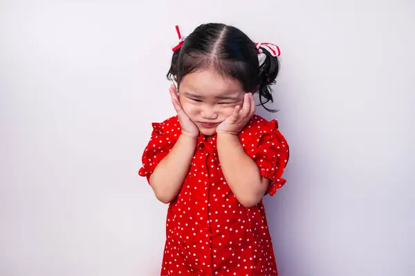 Stock image Asian toddler girl with both hands on cheeks, wearing a red polka dot dress and red and white ribbons on hair isolated on white background.