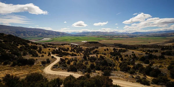 Backcountry nature panorama of road winding through dry rural landscape and green agriculture fields in background near Bendigo Central Otago South Island New Zealand