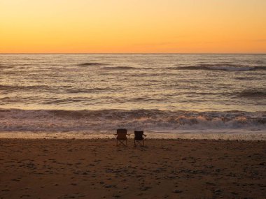 Gillespies Sahili 'nde iki boş kamp sandalyesi ve günbatımında arka planda okyanus dalgaları var. Batı Yakası Güney Adası Yeni Zelanda