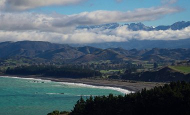 Yeni Zelanda doğa manzarası, turkuaz mavi Pasifik okyanus suyu, siyah kum plajı, yuvarlanan tepeler ve Kaikoura Canterbury Güney Adası 'ndan görülen Güney Alp dağları