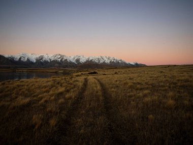 Çimen yol lastik izleri, sakin, sakin, sakin kar saatine yol açıyor. Güney Alpler karla kaplı, Heron Gölü kış günbatımı manzaralı, Ashburton Gölleri Canterbury Güney Adası Yeni Zelanda.