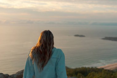 Unrecognizable young woman sitting watching the sea at sunset on Donios beach from the viewpoint of Monteventoso. Ferrol, Galicia.