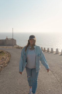 Güneş gözlüklü genç bir kadın, Nazare 'deki San Miguel kalesindeki sörf müzesinden mutlu bir şekilde yürüyor. Portekiz 'de yaz tatili.