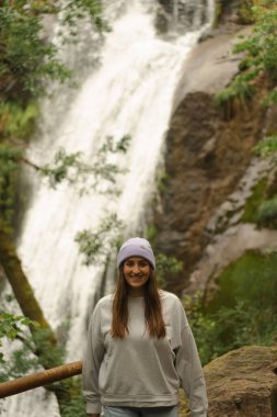 Sıcak giyinmiş genç bir kadın serin bir günde huzurlu bir yürüyüş sırasında Ribasieira şelalesinin önünde gülümsüyor. Bereketli yeşillik onu Porto do Son, Galiçya, İspanya 'da sarmış..