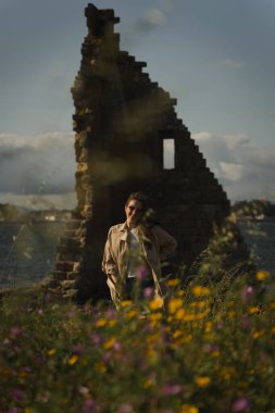 Vertical image of a woman in a trench coat walking towards the tower of San Sadurnino. Coloured flowers in the foreground. Sunset in Cambados, Pontevedra. clipart