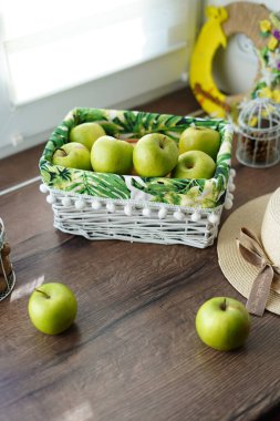A white wicker basket with green juicy apples on a wooden table near the window. Next to the basket lies a beige hat and a walnut basket. Sunny day. Beautiful background