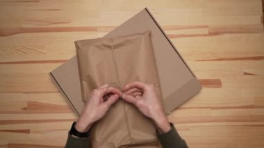 two men's hands pack the package in craft brown paper and place it in a brown paper box against a wooden table. Delivery man hands with cardboard box mockup. Top view