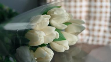 Close-up of a bouquet of white tulips. White tulips lie in a decorative white wicker basket on a wooden table near the window