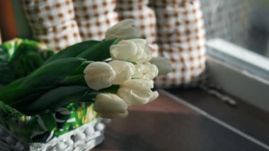 White tulips lie in a decorative white wicker basket on a wooden table near the window against a brown and white decorative cushion