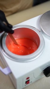 Hot orange wax used for hair removal. Close-up of depilation master hands in black protective gloves holds spatula with melted wax. Device for melting wax. Vertical Screen Orientation Video 9:16