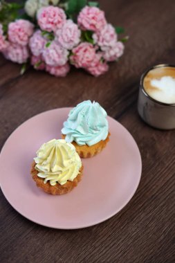 Two delicious, Sweet cream Cakes on a pink plate with yellow and blue cream sit on a wooden table. A cup of cappuccino with wooden Capcakes on the background. Close-up. Copy space