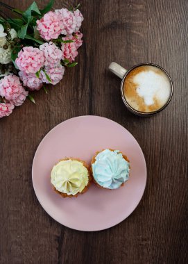 A top view of Two yellow and blue Cream Capcakes on a pink plate sit on a Wooden Table. A beige cup of Cappuccino sits on the table. Near a plate of cakes lie beautiful pink and white flowers.