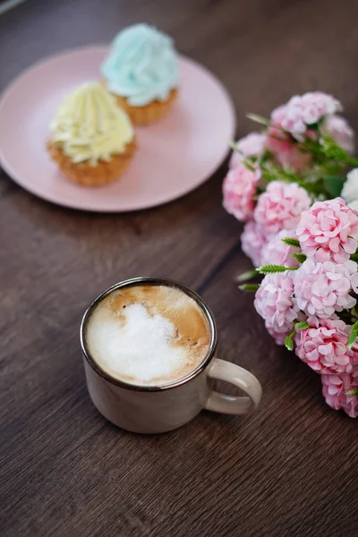 A beige up with appuccino and wo delicious, sweet cream akes on a pink plate with yellow and blue cream sit on a wooden table. A cup of cappuccino with apcakes on the wooden background.