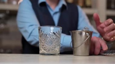 Bartender with steel tongs Puts Ice in Whisky Glass. Elegant bartender in blue shirt and black vest stands behind Bar counter and makes Cocktail. Close up