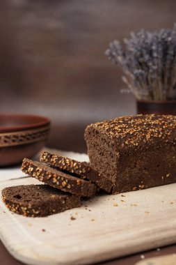 Tahta kesme tahtasının üzerinde kahverengi arka planda dilimlenmiş tahıllı siyah ekmek. Ekmeğin yanında bir kil tabak, çatal, kaşık ve beyaz pamuk masa örtüsü var. Dikey çekim