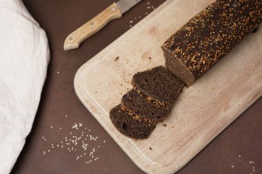 Tahta bir kesme tahtasının üzerinde kahverengi bir arka planda dilimlenmiş tahıllı bir Kara Ekmek Manzarası. Bir bıçak ve beyaz bir pamuk masa örtüsü Ekmeğin yanında yatar