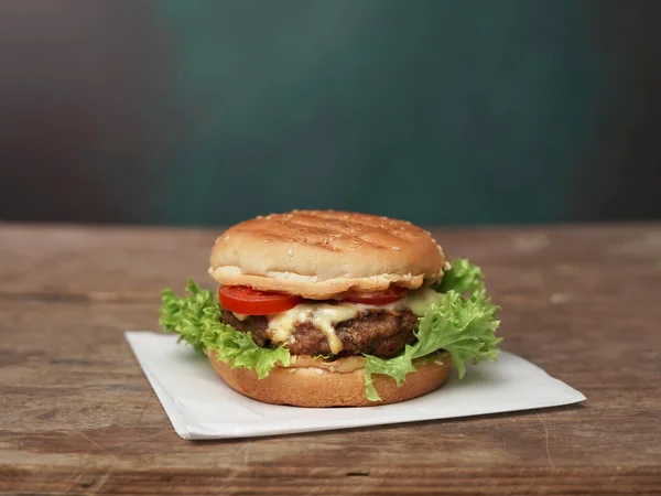 Big Burger lies on craft white paper against wooden table. A Juicy green Salad leaf and a red Tomato lie near the Burger