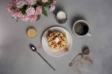Lezzetli waffle tabağı, karamel sosu, kahve fincanı, süt, tatlı kaşığı, süzgeç, gri beton arka planda pembe çiçekler.