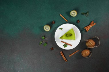 Yeşil arka planda beyaz tabakta Kiwi soslu ev yapımı cheesecake. Bir dilim kivi, tarçın çubuğu ve nane yapraklarıyla süslenmiş. Üst görünüm