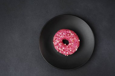 Siyah tabakta renkli şekerlemeler olan pembe jöleli bir çöreğin en üst görüntüsü. Siyah arka planda pembe çörek.