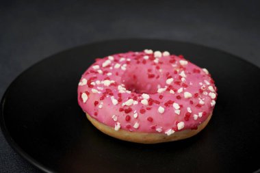 Siyah bir tabakta renkli serpiştirilmiş Pembe Jöleli Donut. Siyah arka planda pembe çörek.
