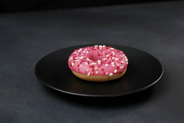 stock image Pink glazed Doughnut with colorful sprinkles on a black plate. Pink donut on a black background