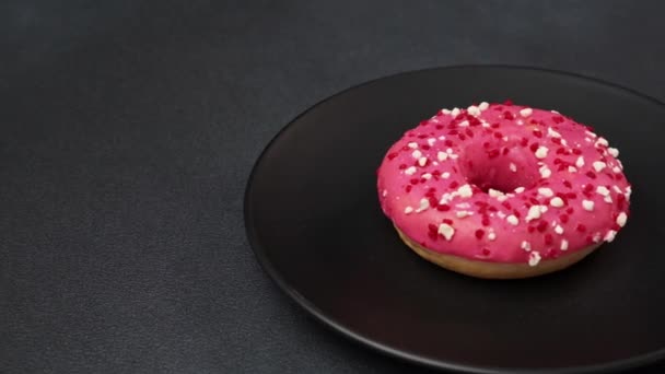粉红釉面甜甜圈的特写镜头 黑色盘子上有五颜六色的洒水 粉红甜甜圈 黑色背景 — 图库视频影像