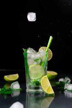 Buz küplerini siyah arka planda bir bardak Mojito 'ya düşürmek. Mojito Kokteylinin limon, buz küpleri ve yeşil nane yapraklarıyla tazelenmesi