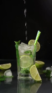 Siyah arka planda bir bardak Mojito 'ya akan bir su. Mojito kokteylini limon, buz küpleri ve yeşil nane yapraklarıyla tazeliyorum. Ağır çekim 120fps. Dikey video