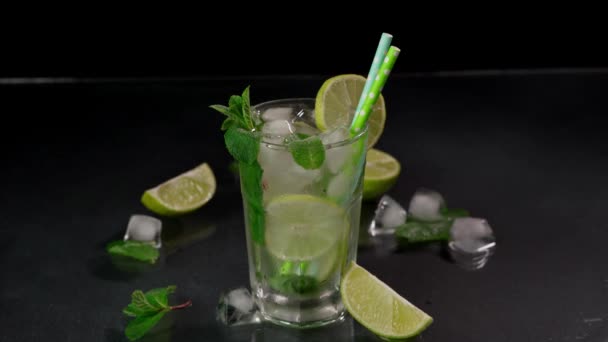黒を背景にライム ラム酒 緑のミントが入ったモヒートのグラスにアイスキューブを入れるバーテンダーの手のクローズアップ — ストック動画
