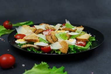Tavuklu Sezar Salatası, marul yaprakları, kiraz domatesleri, siyah bir tabakta rendelenmiş parmesan.