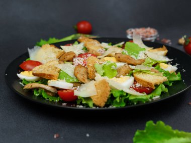 Tavuklu Sezar Salatası, marul yaprakları, kiraz domatesleri, siyah bir tabakta rendelenmiş parmesan.