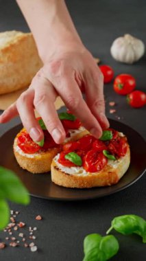 Ricotto peyniri ve vişneli domates fesleğen, siyah arka planda sarımsak. Adamın eli ricotto ve domates bruschetta 'yı fesleğenle süslüyor. Dikey video