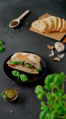 Jambonlu Ciabatta Sandviçi, Pesto sosu, Domates, siyah arka planda Basil. Yiyecekler. Yemek arkaplanı. Dikey video
