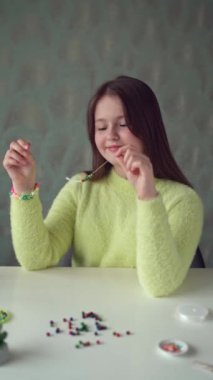 Uzun saçlı ve sarı kazaklı küçük bir kız boncuklu mücevherler örüyor. Dikey video