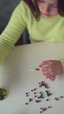 Uzun saçlı ve sarı kazaklı küçük bir kız boncuklu mücevherler örüyor. Yakın plan. Dikey video