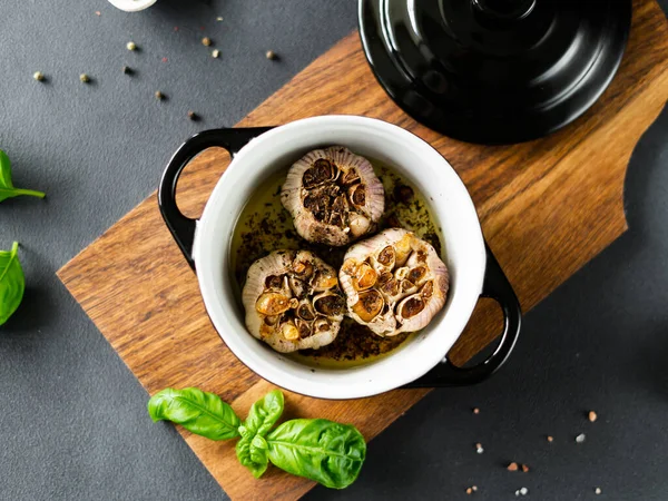 stock image Garlic bulbs Baked with olive oil and spices in black dish. Garlic roasted, black pepper and basil on wooden board. Top view