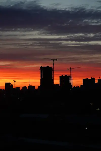 Sunset against the backdrop of a large construction site with several working cranes and unfinished high-rise buildings. Sunset with orange and red sky in cityscape with silhouettes of buildings