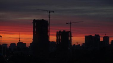 Gün batımını, çok sayıda çalışan vinç ve tamamlanmamış yüksek binalarla birlikte büyük bir inşaat sahasının arka planına karşı. Şehir manzarasında turuncu ve kırmızı gökyüzü olan gün batımı binaların siluetleriyle