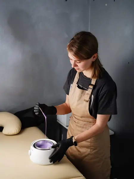 Hot violet wax used for hair removal. Close-up of depilation master hands in black protective gloves holds spatula with melted wax. Device for melting wax