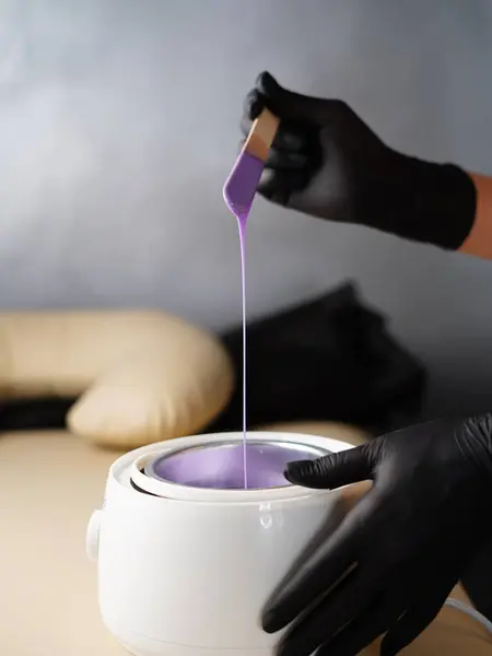 Hot violet wax used for hair removal. Close-up of depilation master hands in black protective gloves holds spatula with melted wax. Device for melting wax