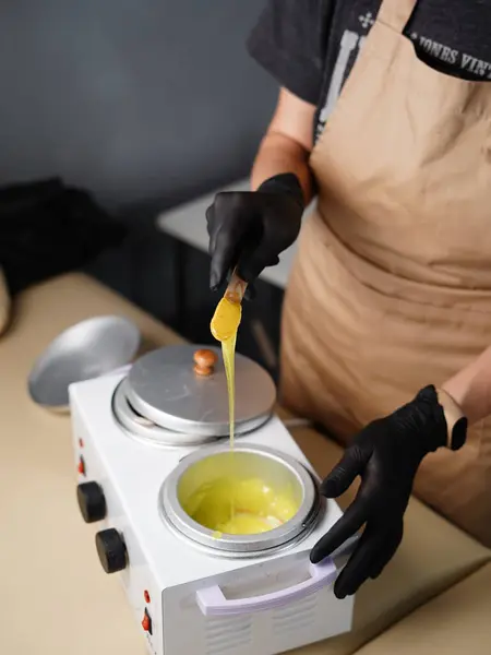 Hot gold wax used for hair removal. Close-up of depilation master hands in black protective gloves holds spatula with melted wax. Device for melting wax