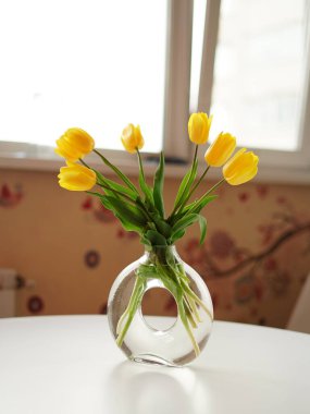 Sarı lale buketi şeffaf cam vazoda beyaz bir masada duruyor.