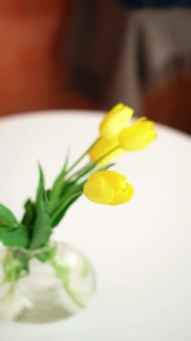 Sarı lale buketi şeffaf cam vazoda beyaz bir masada duruyor.