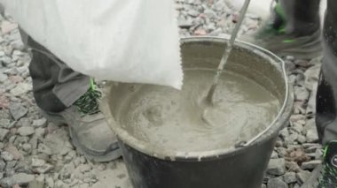 İnşaatçı, çimentoyu siyah kovaya karıştırıyor. Harekete odaklanıyor. Arka plan çakıl taşından oluşuyor, ve kişi iş pantolonu ve sağlam iş ayakkabıları giyiyor, bu da bir yapıyı işaret ediyor.