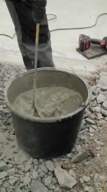 İnşaatçı, çimentoyu siyah kovaya karıştırıyor. Harekete odaklanıyor. Arka plan çakıl taşından oluşuyor, ve kişi iş pantolonu ve sağlam iş ayakkabıları giyiyor, bu da bir yapıyı işaret ediyor.