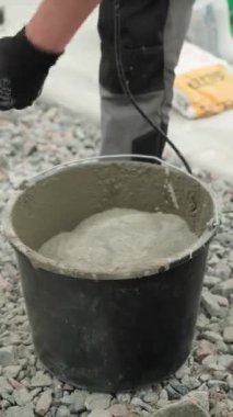 İnşaatçı, çimentoyu siyah kovaya karıştırıyor. Harekete odaklanıyor. Arka plan çakıl taşından oluşuyor, ve kişi iş pantolonu ve sağlam iş ayakkabıları giyiyor, bu da bir yapıyı işaret ediyor.